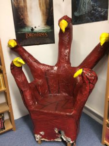 Ein Stuhl in Form einer großen, roten Klaue mit gelben Fingernägeln. Er befindet sich in einer Bibliothek.